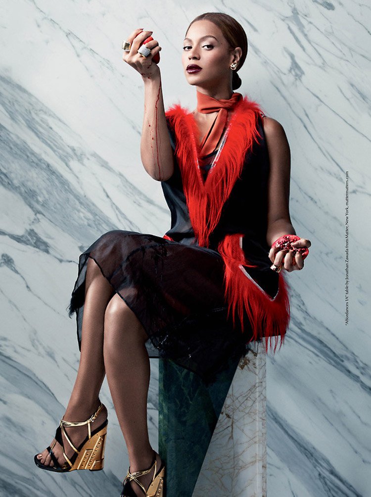 Beyonce seated in a studio setting, wearing a black dress with a red fur vest, holding fruit, and showcasing gold platform heels.