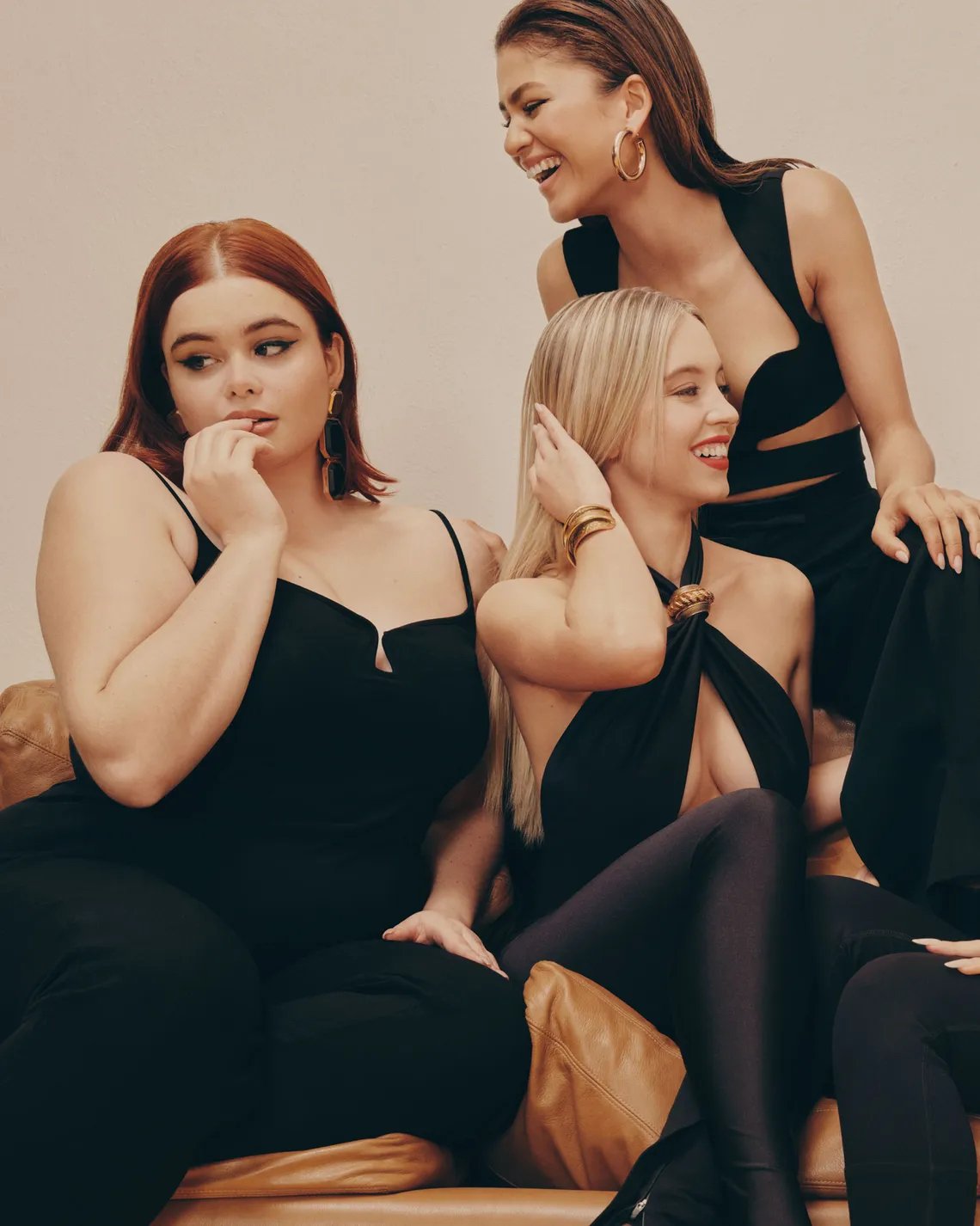 Three women wearing stylish black outfits pose indoors on a brown leather couch; one woman is laughing in this studio shot.