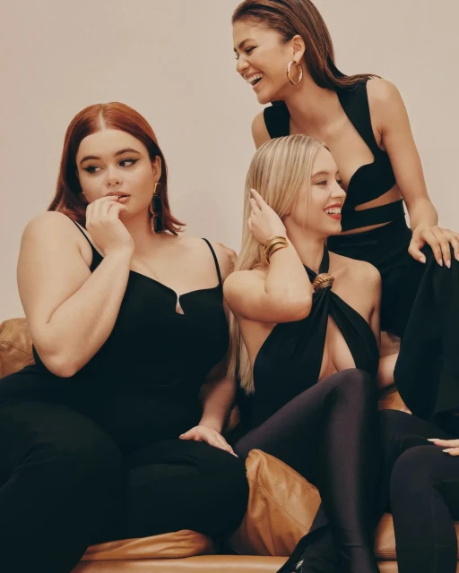 Three women wearing stylish black outfits pose indoors on a brown leather couch; one woman is laughing in this studio shot.