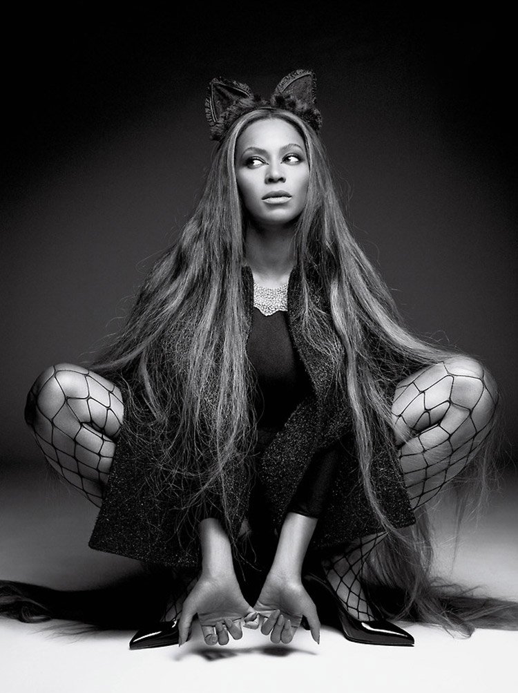 Black and white portrait of a woman with long hair, fishnet stockings, cat ears, and high heels crouching against a dark background.
