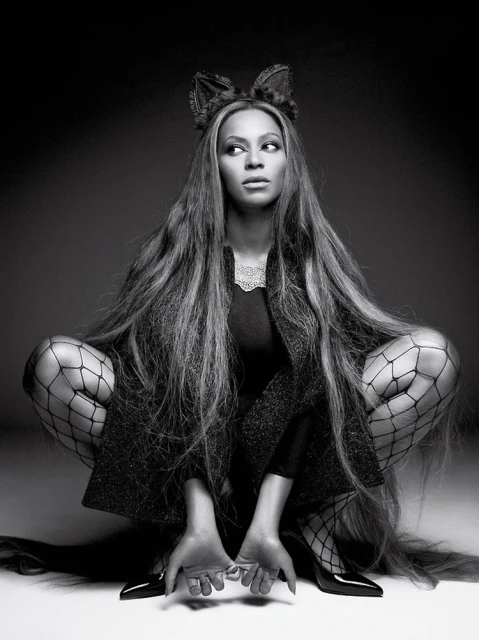 Black and white portrait of a woman with long hair, fishnet stockings, cat ears, and high heels crouching against a dark background.
