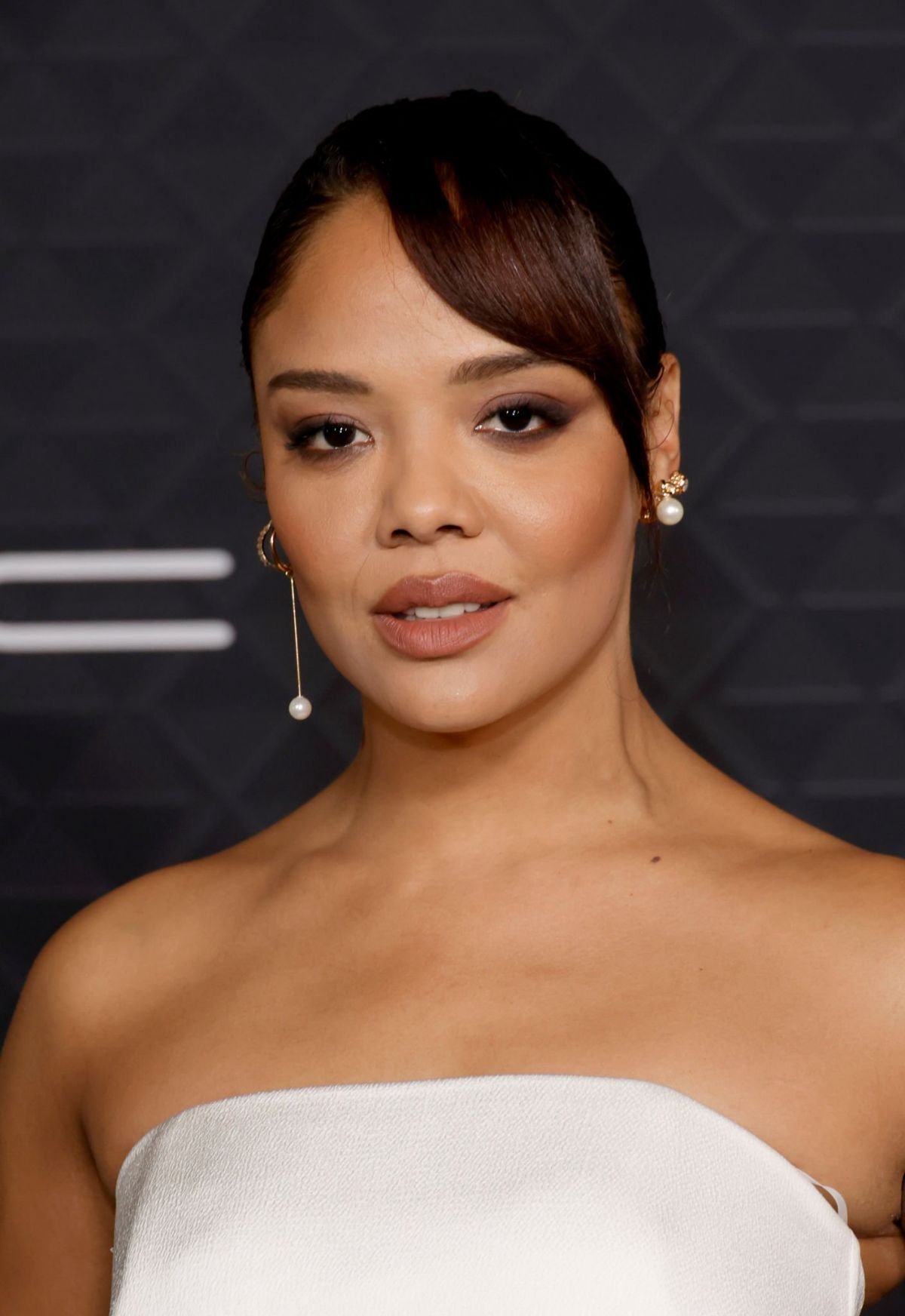 Close-up shot of a woman with dark hair and fair skin wearing a white strapless top, pearl earrings, and minimal makeup against a grey background.