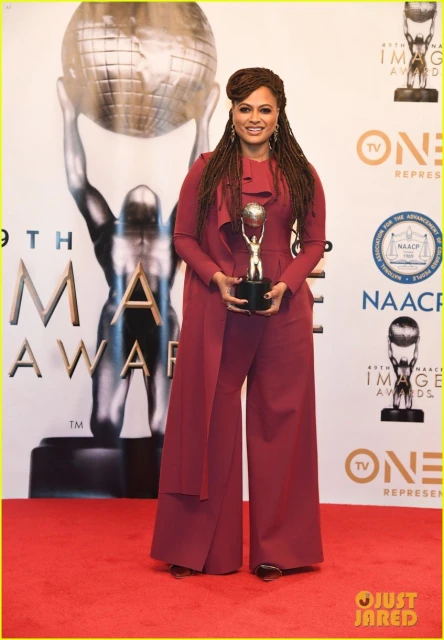 Ava DuVernay at the 45th NAACP Image Awards, holding an award in a maroon jumpsuit in front of the awards backdrop and red carpet.