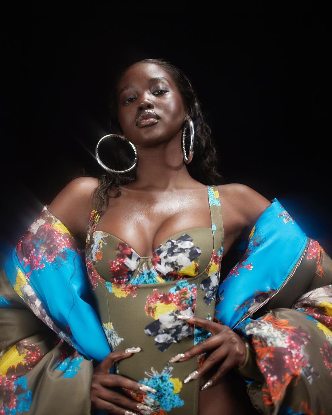 Portrait of an African American woman wearing a floral bodysuit and a floral puffer jacket over her shoulders in front of a black background.