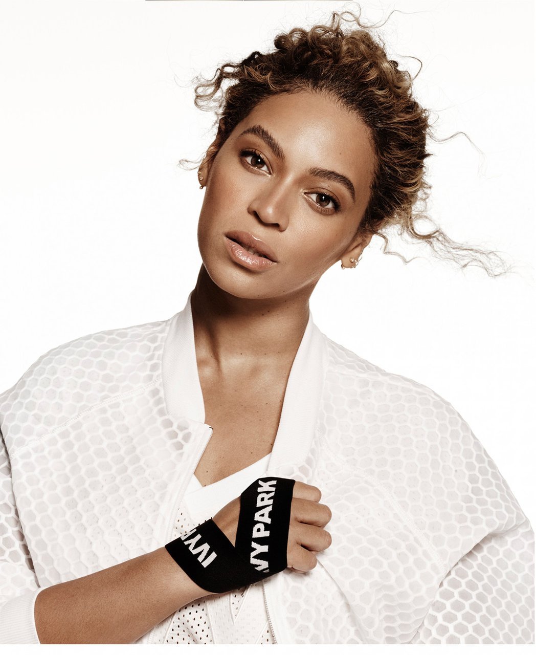 Beyoncé poses in a white sports jacket with 'Ivy Park' branded wristbands in a studio, showcasing fashion and fitness.