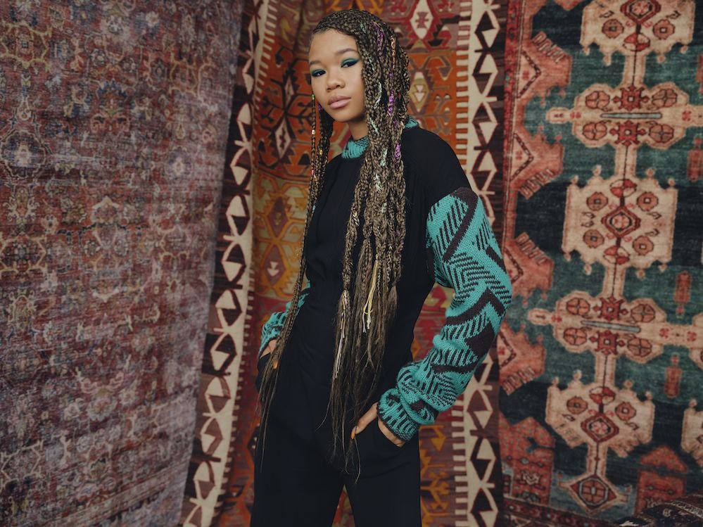 Young woman with braided hair in black and green patterned sweater standing against a backdrop of ornate rugs in an indoor setting.