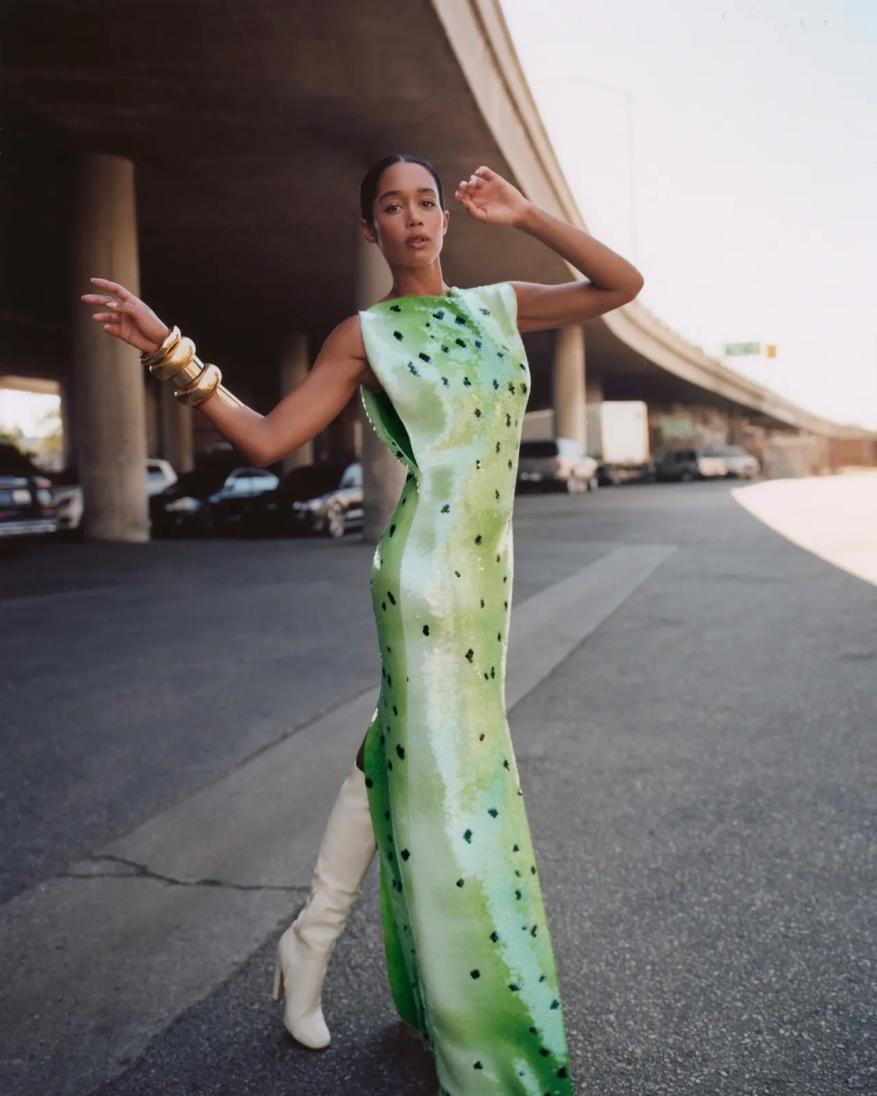 Adult female fashion model poses in a green spotted evening gown with beige thigh high boots on a city street under a highway overpass.