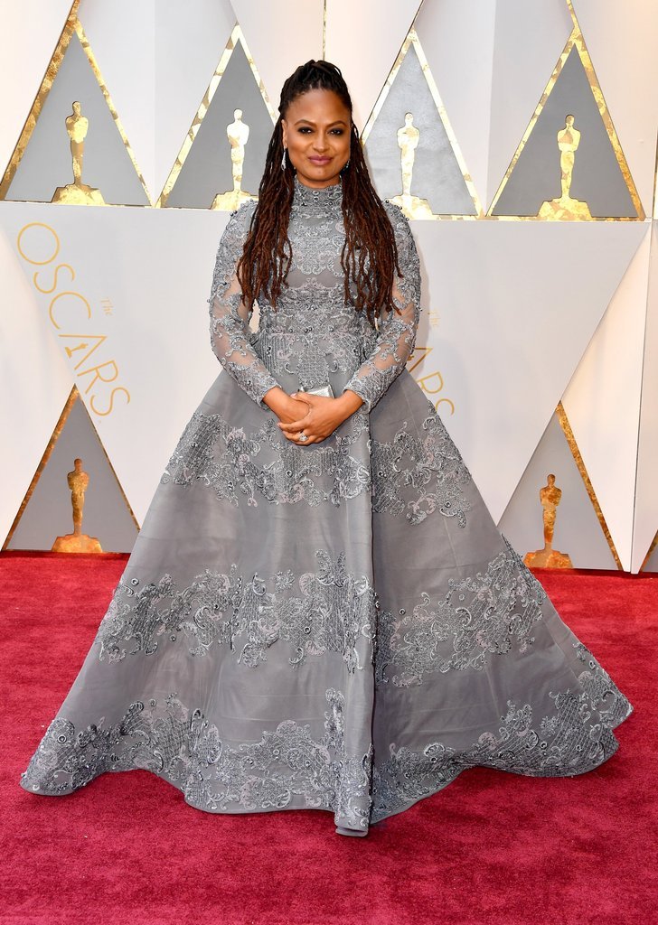 A woman poses on a red carpet wearing a gray gown with lace details, long sleeves, and a high neckline for a formal event.