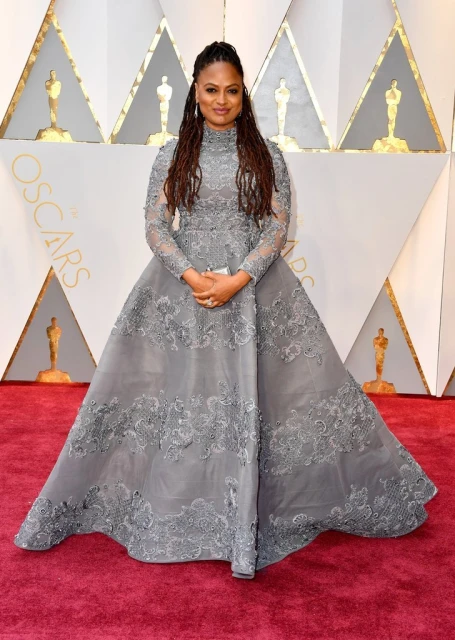 A woman poses on a red carpet wearing a gray gown with lace details, long sleeves, and a high neckline for a formal event.