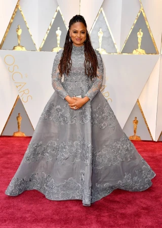 A woman poses on a red carpet wearing a gray gown with lace details, long sleeves, and a high neckline for a formal event.