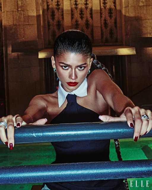 Zendaya poses wearing a halter top and black tie with a white collared shirt in front of metal bars against an indoor background.