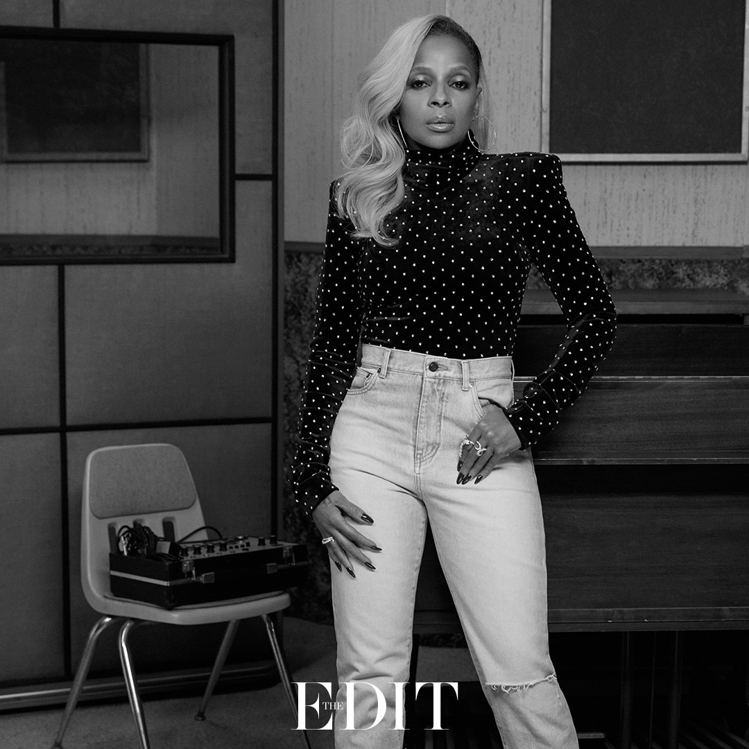 Mary J. Blige in a black and white portrait, wearing a black studded top and light jeans, posing indoors with fair skin and blonde hair.