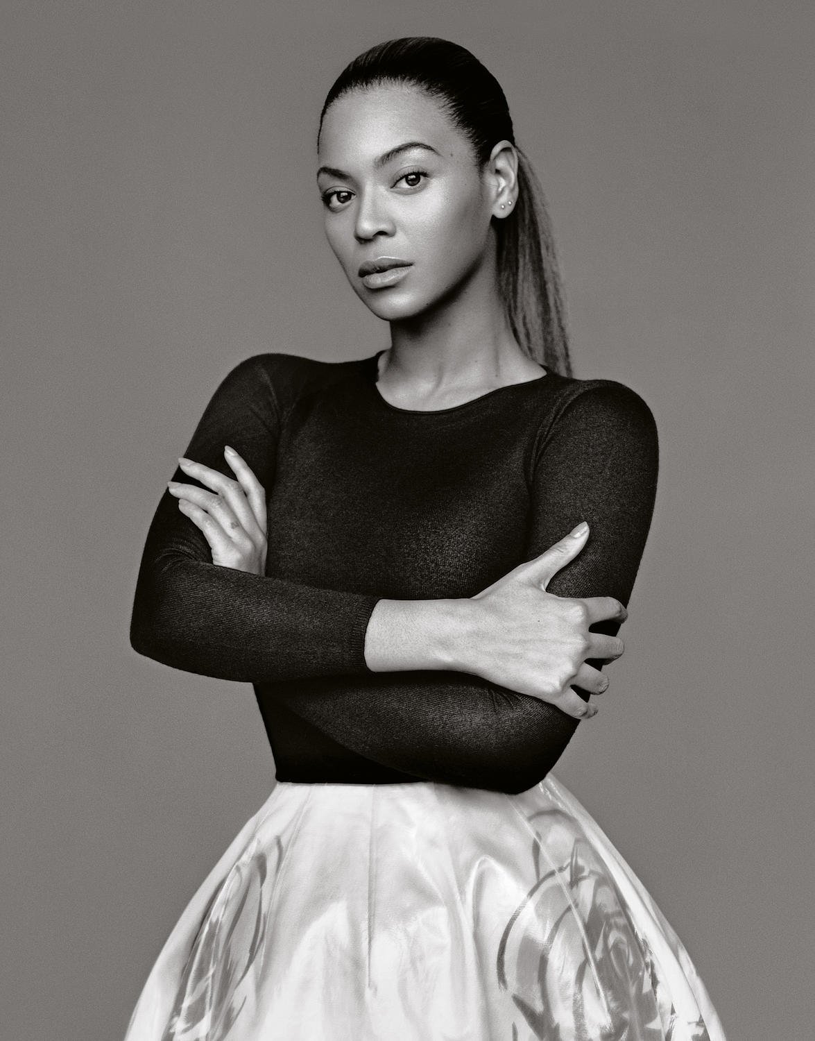Portrait of Beyonce with arms crossed, wearing a black top and white skirt, captured in a monochrome studio shot against a grey backdrop.