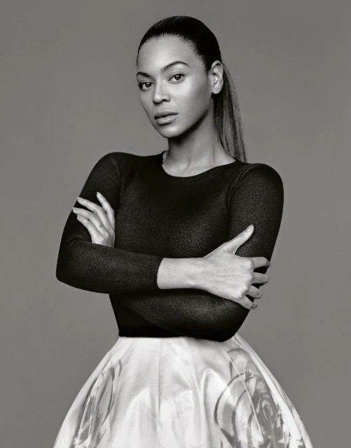 Portrait of Beyonce with arms crossed, wearing a black top and white skirt, captured in a monochrome studio shot against a grey backdrop.
