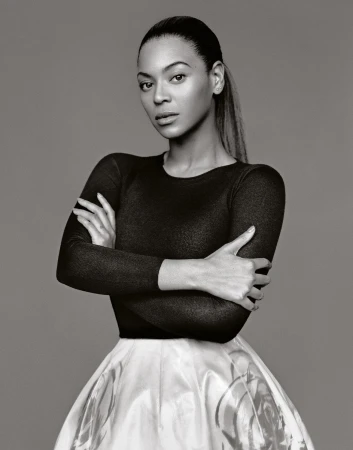 Portrait of Beyonce with arms crossed, wearing a black top and white skirt, captured in a monochrome studio shot against a grey backdrop.