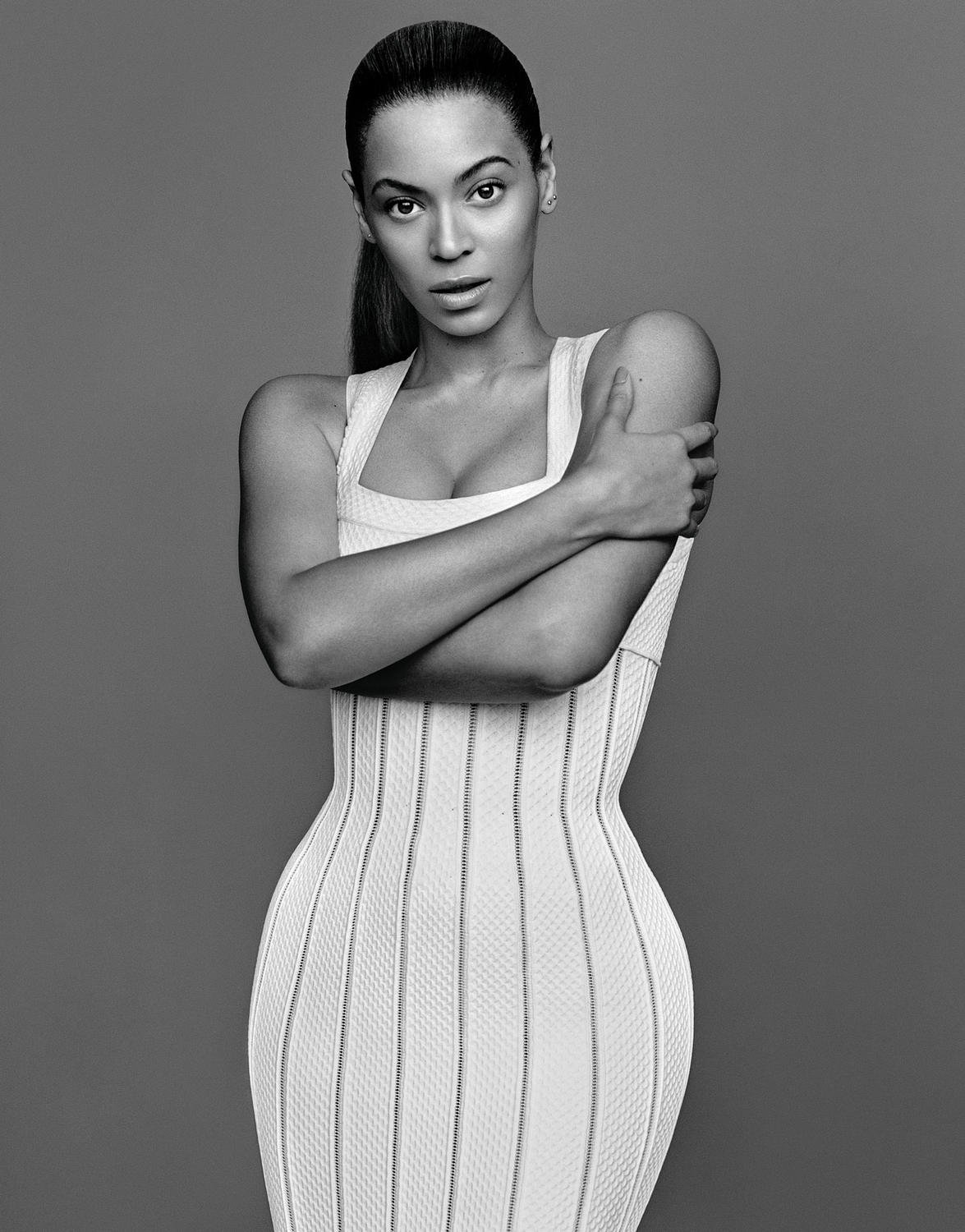 Monochrome studio portrait of Beyonce with arms crossed, wearing a tight white dress against a solid grey backdrop, looking at camera.