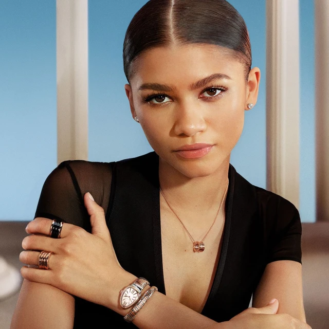 A brunette woman poses in a studio wearing a sheer black top and Bulgari jewelry against a background with blue and white pillars.