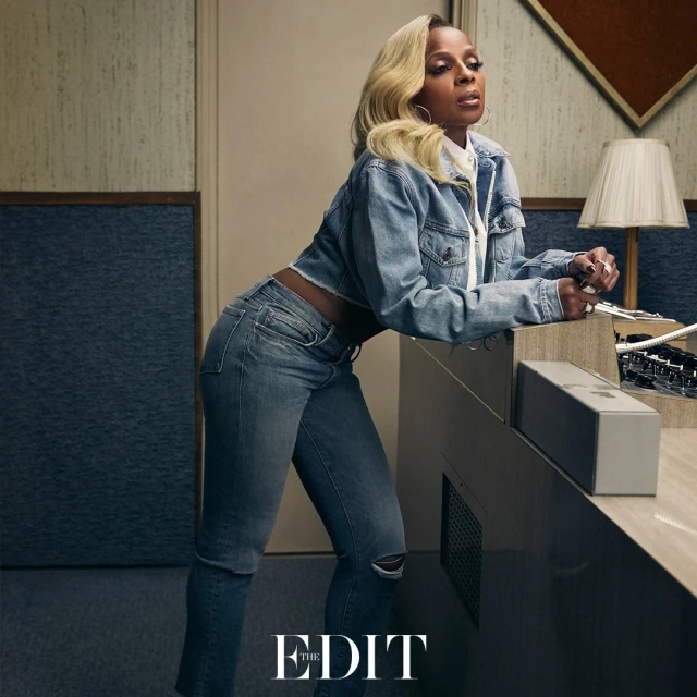 Blonde-haired woman with a denim jacket and jeans, leaning on a counter with a mixing board, in a studio setting, with warm lighting.