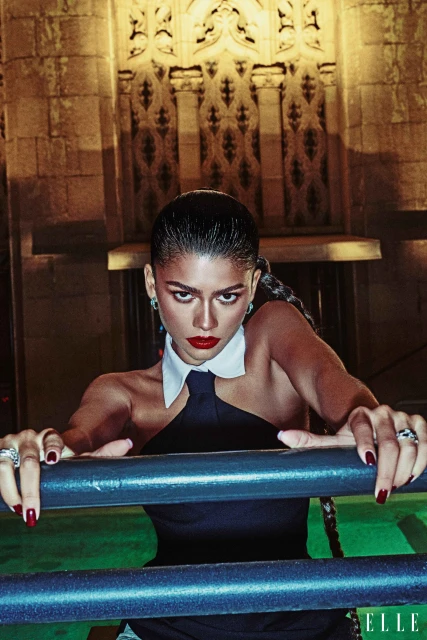 Portrait of adult female posing with red lipstick, dark top, and white collar leaning against a blue metal fence with an ornate building.