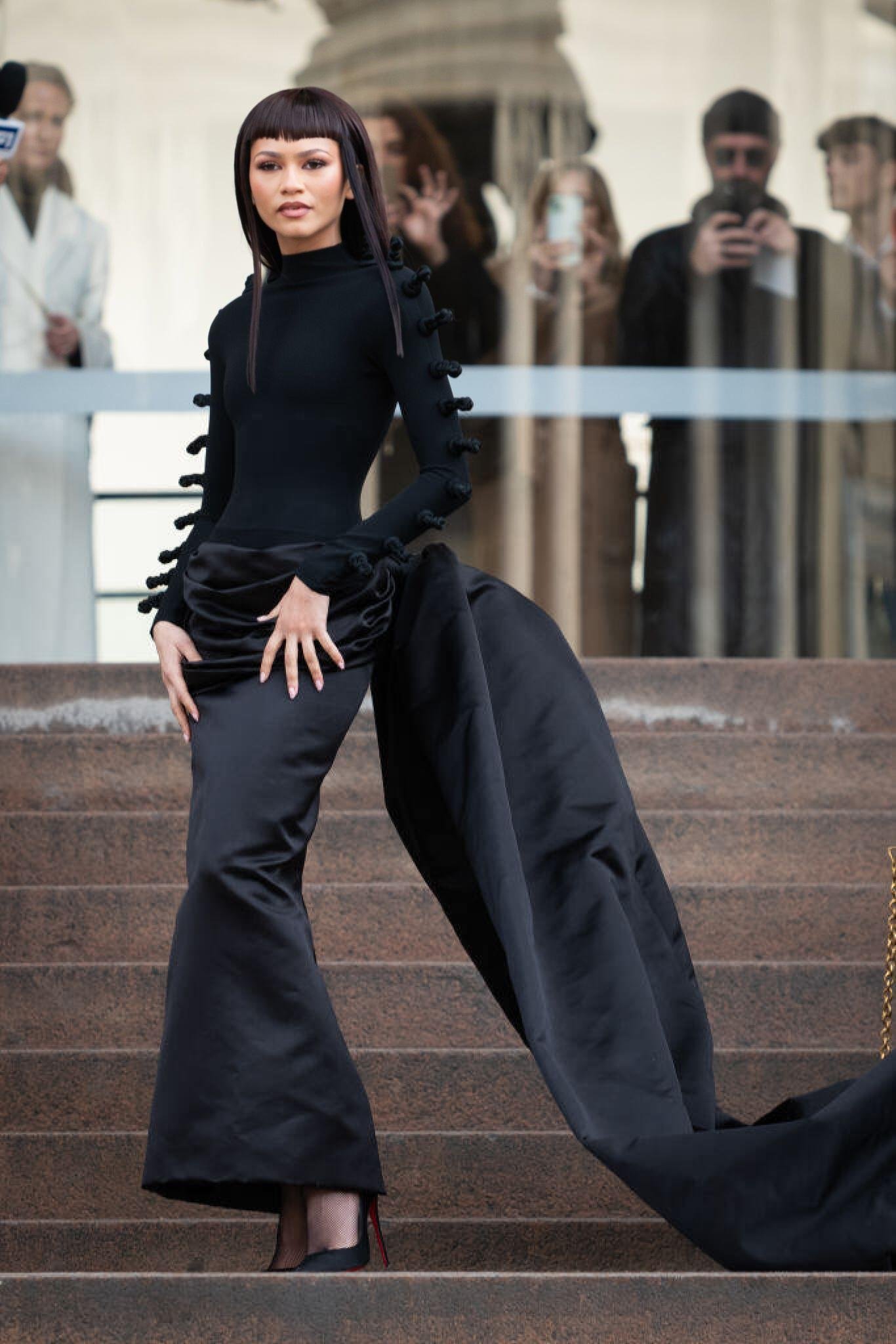 Zendaya strikes a confident pose on stone steps, wearing a dramatic black gown with long train; onlookers blur in background.