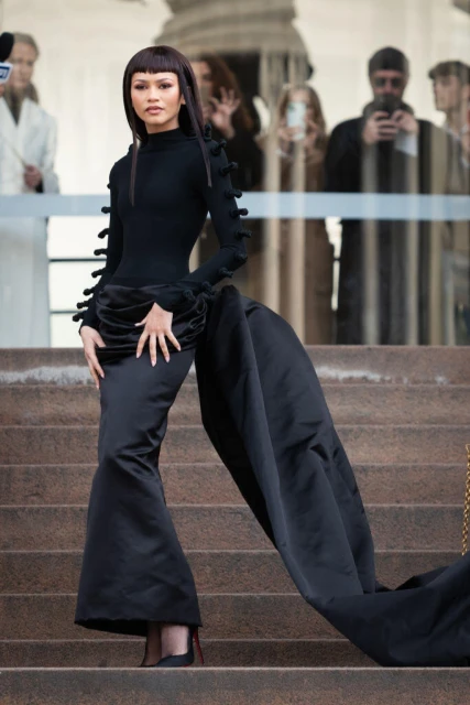Zendaya strikes a confident pose on stone steps, wearing a dramatic black gown with long train; onlookers blur in background.