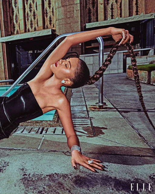 Glamorous woman with braided ponytail, jewelry, and strapless corset poses poolside on a textured surface in editorial fashion shot.