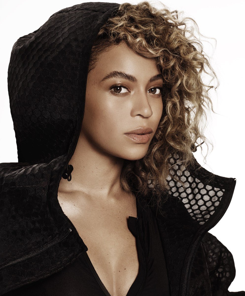 Close-up portrait of a woman with curly hair wearing a hooded jacket, set against a white background. Beauty and fashion aesthetic.