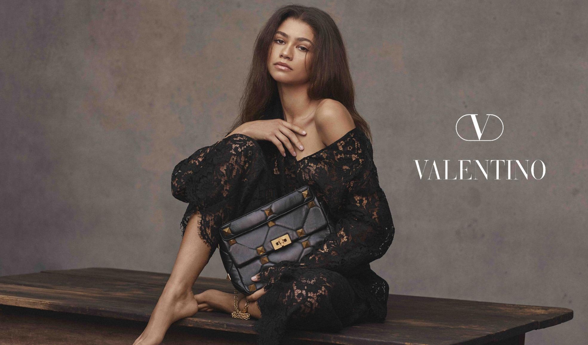 Zendaya poses with a Valentino Garavani studded Roman Stud top-handle bag wearing a black lace dress while seated on a wooden table.
