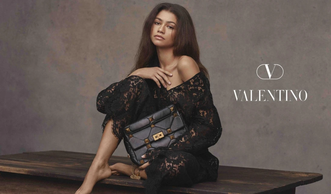 Zendaya poses with a Valentino Garavani studded Roman Stud top-handle bag wearing a black lace dress while seated on a wooden table.