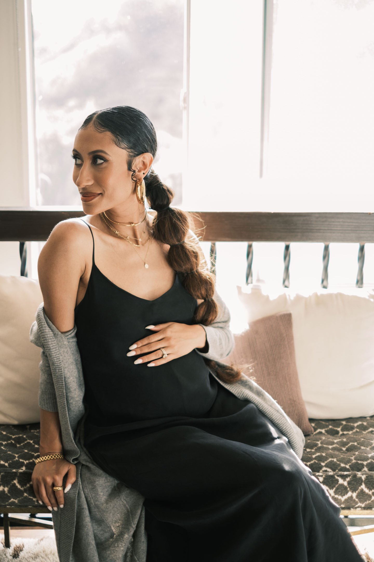 A woman in a black slip dress and grey cardigan sits indoors with her hand on her stomach near a bright window.