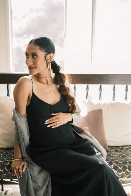 A woman in a black slip dress and grey cardigan sits indoors with her hand on her stomach near a bright window.