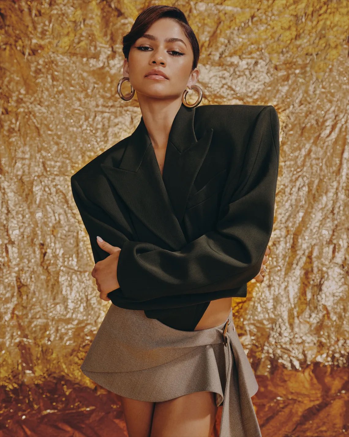 Zendaya wearing a structured black blazer and grey wrap skirt with large hoop earrings against a textured metallic gold backdrop