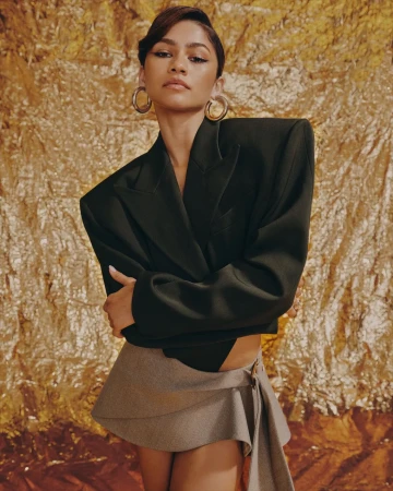 Zendaya wearing a structured black blazer and grey wrap skirt with large hoop earrings against a textured metallic gold backdrop