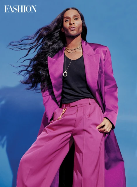 Fashionable adult male model with long dark hair poses in a purple satin suit against a blue gradient background in a studio fashion shot.