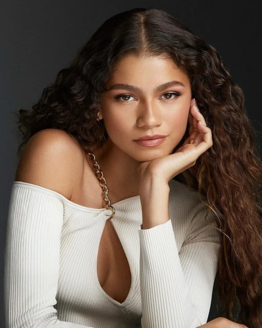 Portrait of a woman with long curly hair wearing a white ribbed top with cut out detail, posing in a photo studio.
