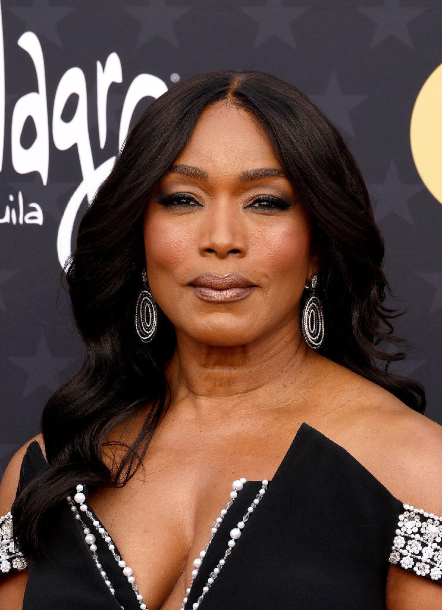 Close-up portrait of Angela Bassett in a black dress, smiling on a red carpet with decorative earrings and black wavy hair.