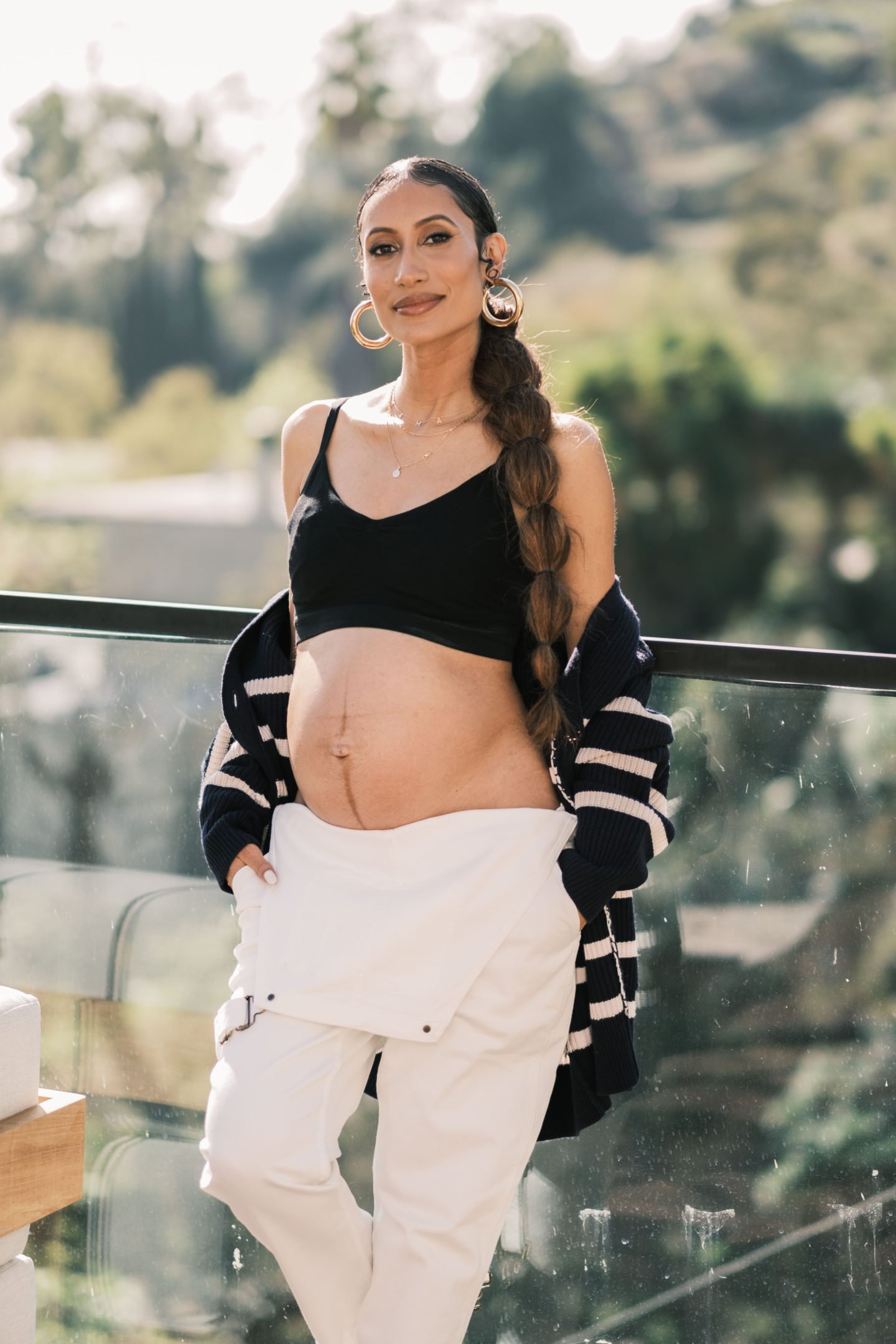 Pregnant woman with long braid, wearing a black top, white pants and a striped cardigan poses outdoors on a sunny day.
