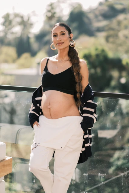 Pregnant woman with long braid, wearing a black top, white pants and a striped cardigan poses outdoors on a sunny day.