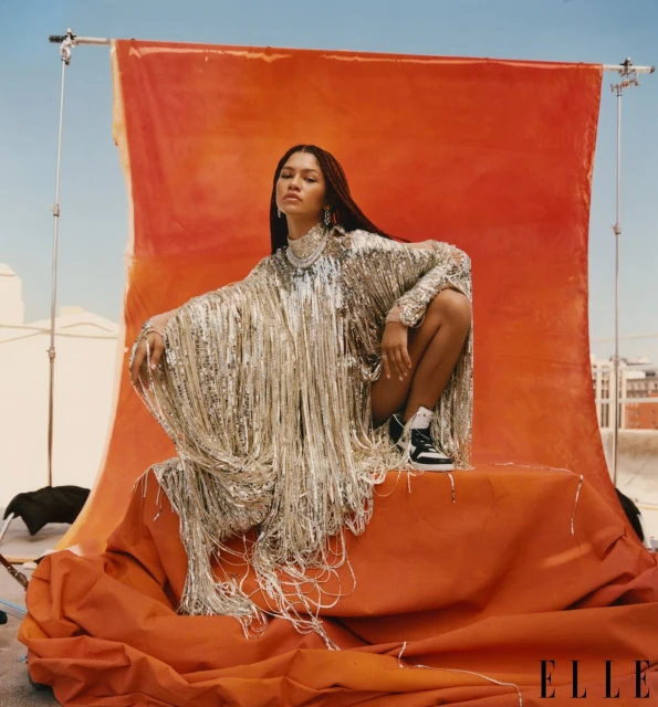 Zendaya sits on orange fabric in a silver fringe dress. A backdrop of orange and blue suggests an outdoor fashion shoot.