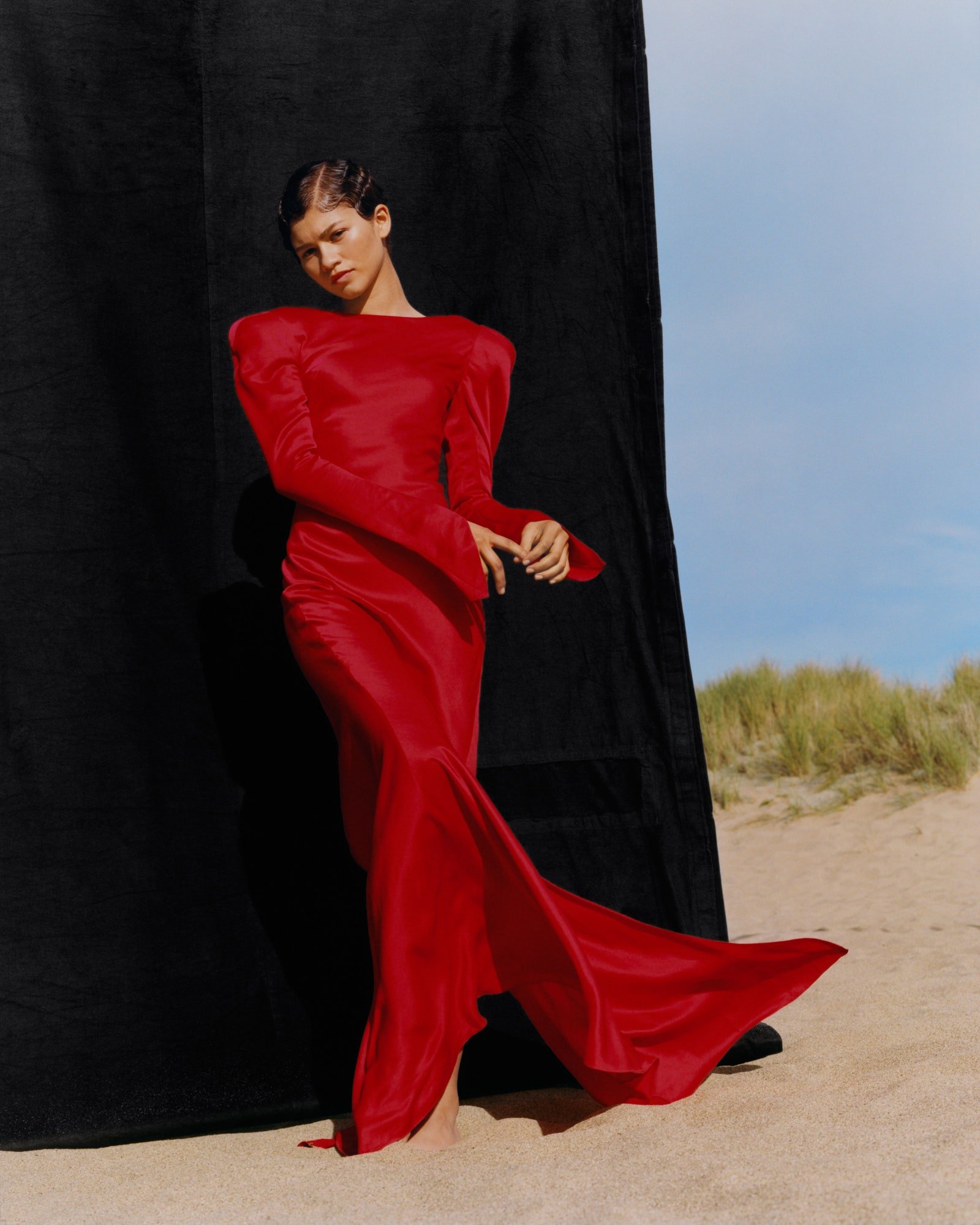 Woman in long-sleeved red gown stands on sandy beach next to dark fabric, posing for a fashion shoot under a bright blue sky.
