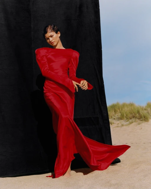 Woman in long-sleeved red gown stands on sandy beach next to dark fabric, posing for a fashion shoot under a bright blue sky.
