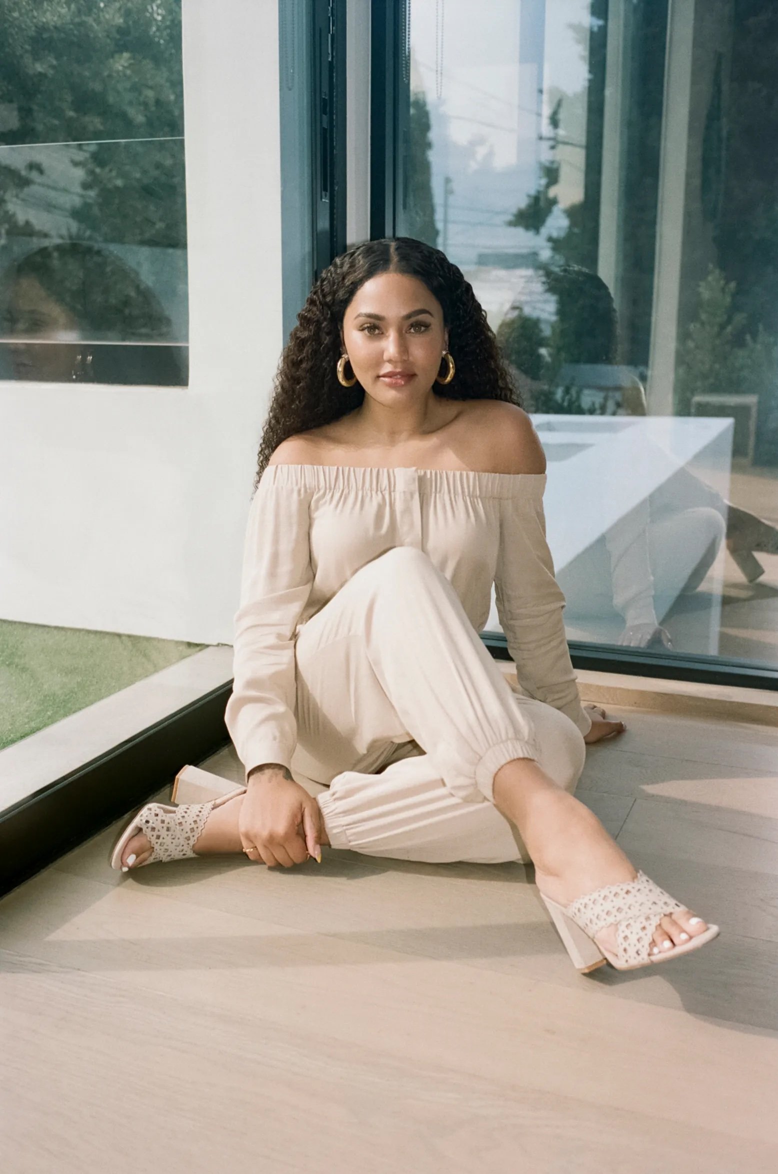 Adult woman in off-the-shoulder top and pants poses while sitting indoors with natural light and a window reflection.
