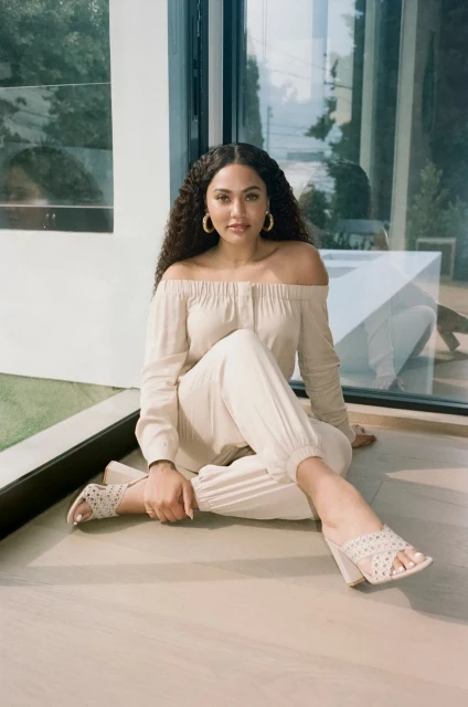 Adult woman in off-the-shoulder top and pants poses while sitting indoors with natural light and a window reflection.