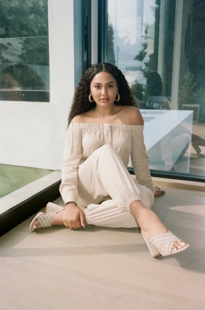 Adult woman in off-the-shoulder top and pants poses while sitting indoors with natural light and a window reflection.