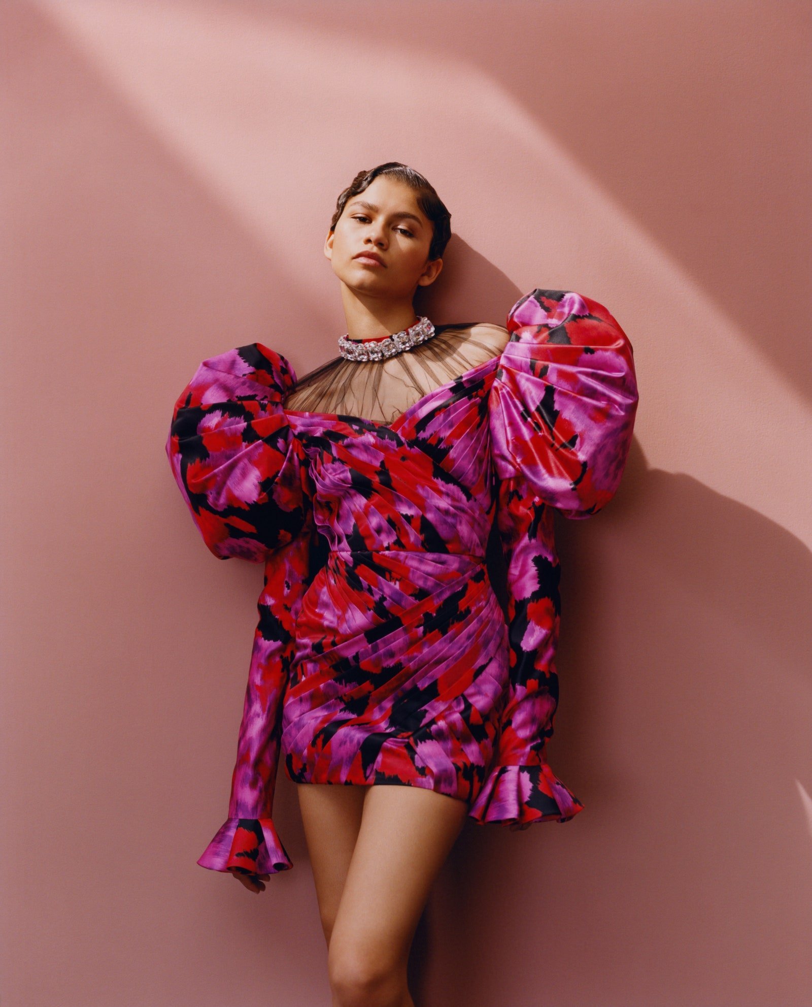 A young adult female wearing a floral mini dress with puffed sleeves and a large jewel necklace poses against a pink backdrop.