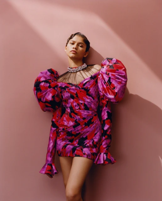 A young adult female wearing a floral mini dress with puffed sleeves and a large jewel necklace poses against a pink backdrop.