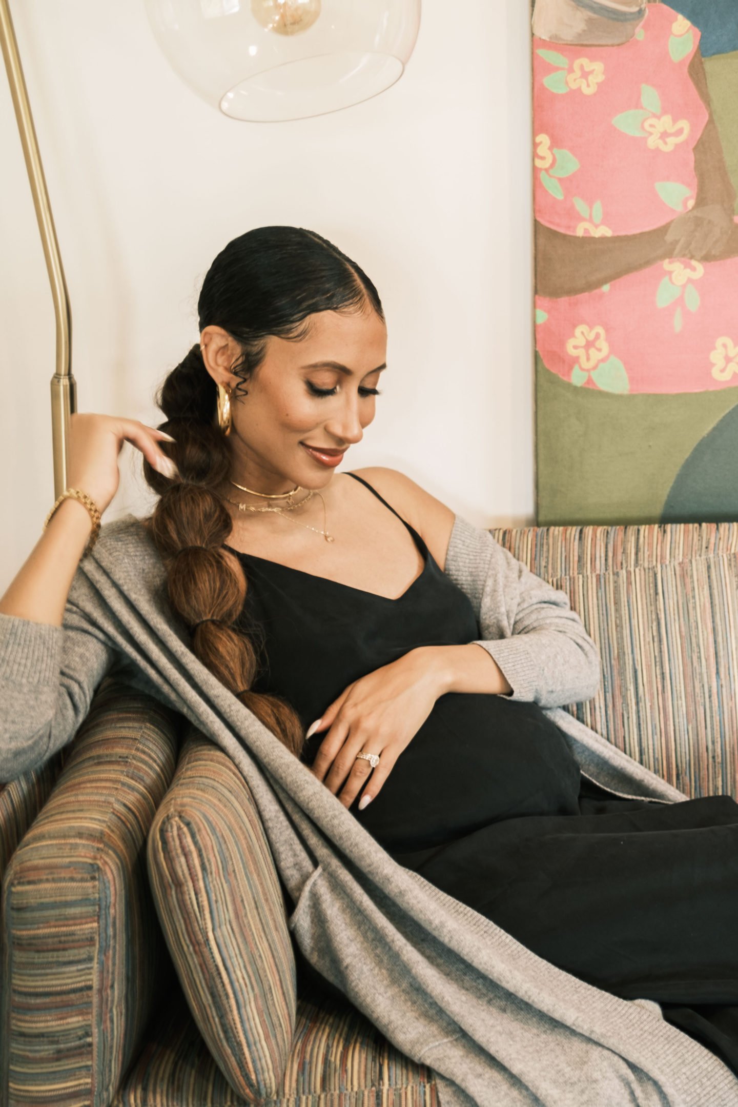 Pregnant woman wearing black dress and grey cardigan sitting on striped chair in natural light with painting in background