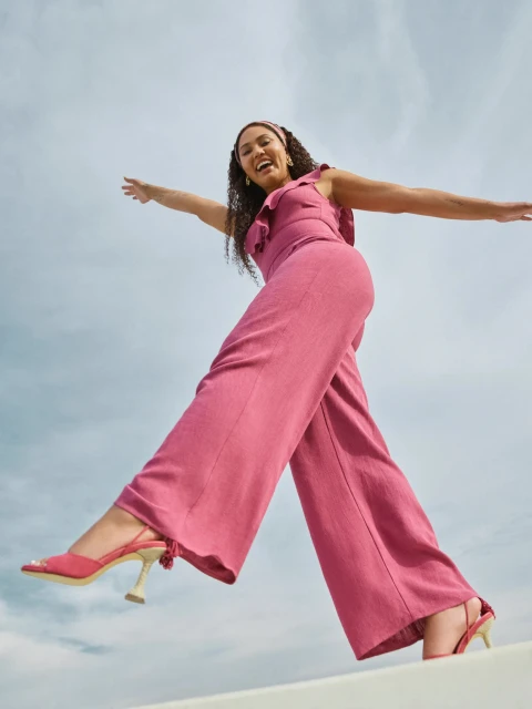 Smiling woman in a pink jumpsuit poses on a white ledge against a cloudy sky, with her arms outstretched for balance and joy.