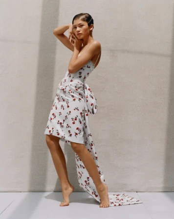 Woman in floral dress with bow poses against a neutral wall, fashion shot with soft lighting and elegant presentation, barefoot.
