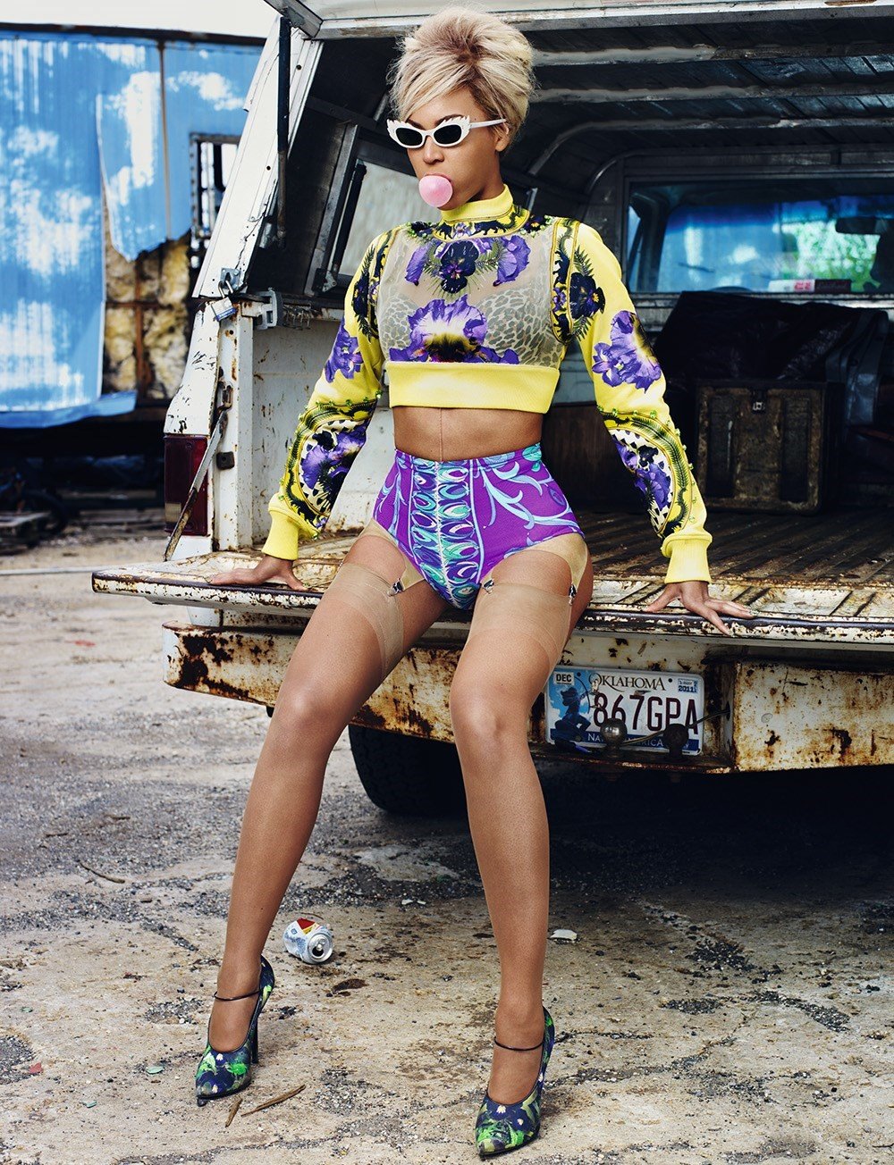 Woman poses on truck wearing yellow crop-top and purple shorts, blowing a bubblegum bubble in an outdoor setting, vintage feel.